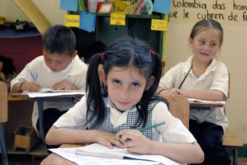 Una niña con uniforme escolar escribe en su cuaderno y mira a la cámara en un aula de Florida, Valle del Cauca.