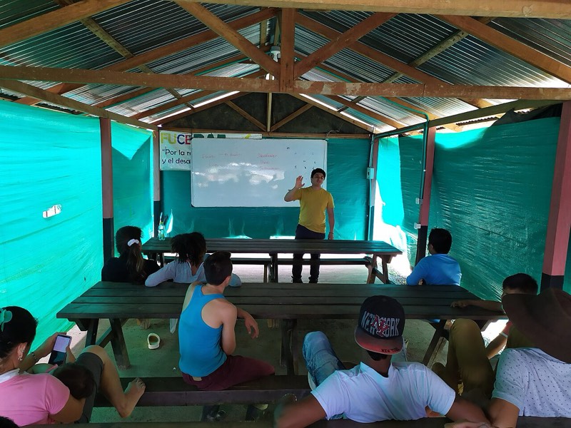 Un joven explica frente a un tablero a otros jóvenes sentados en un aula abierta con techo de zinc.