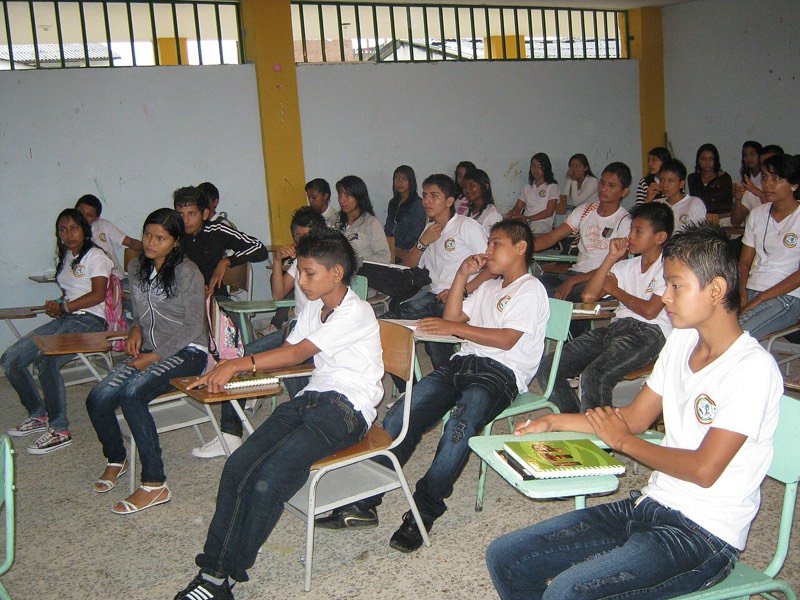 Jóvenes de secundaria con uniforme blanco atienden la clase sentados en pupitres.